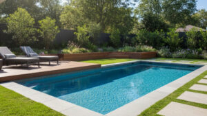 A wide shot of a rectangular plunge pool in a lush, verdant backyard. The pool is flanked by a wooden sun deck with two lounge chairs on one side and a manicured lawn with stepping stones on the other, surrounded by dense trees and flowering plants.